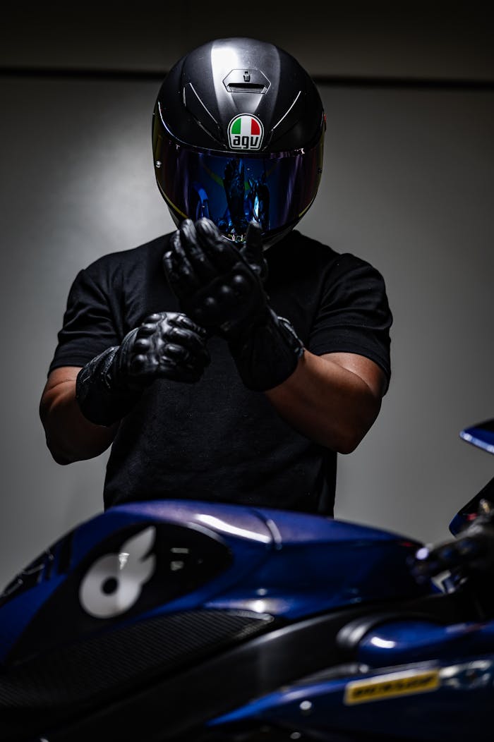 A biker with helmet adjusts gloves beside a motorcycle, in a moody setting.